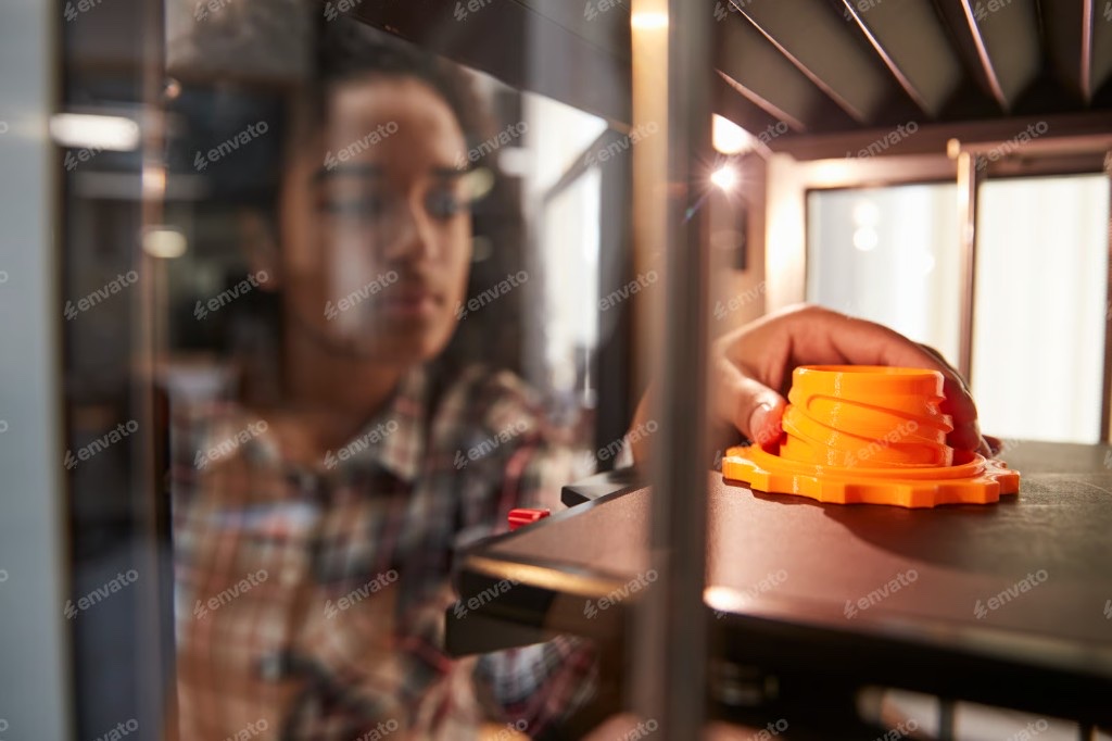 A hand removing an orange 3D-printed part from a printer