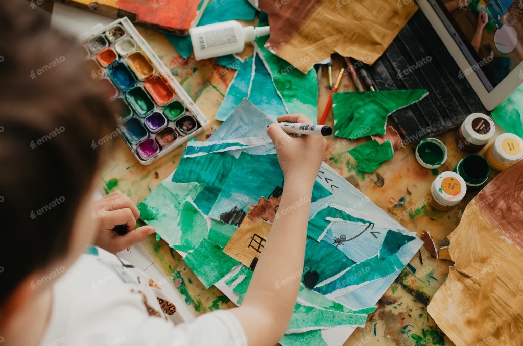 Overhead view of a paint-splattered table with watercolor palette, brushes, and a paper collage