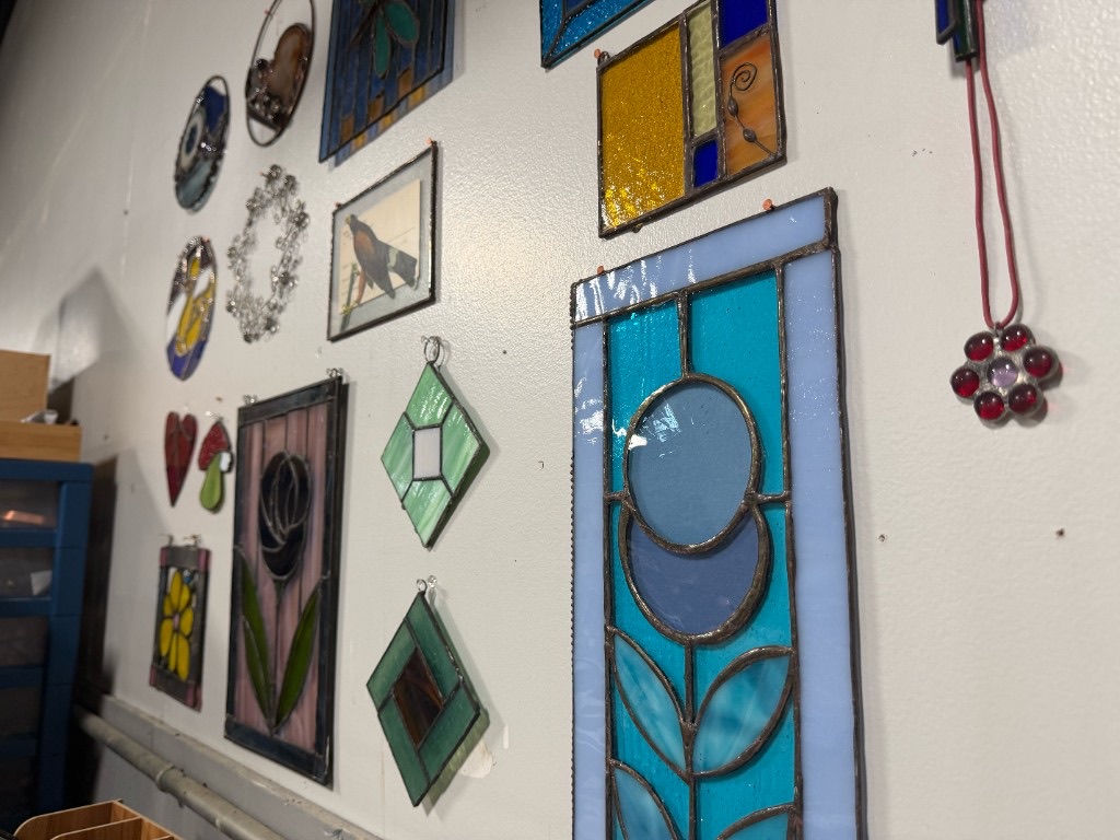 Handmade stained glass ornaments and panels displayed on a white wall